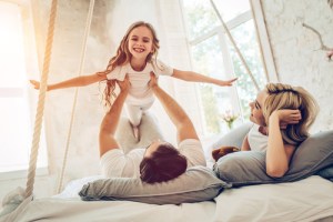 Happy family playing together on bed in bright bedroom, father lifting young daughter in the air while mother watches and smiles