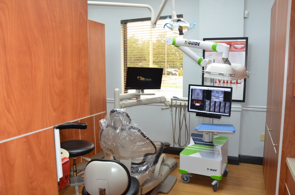 Modern dental treatment room with chair, digital monitors, overhead equipment, and wooden cabinetry