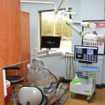 Modern dental treatment room with chair, digital monitors, overhead equipment, and wooden cabinetry