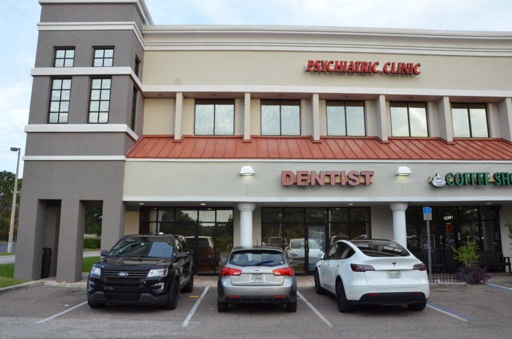 Two-story commercial building with dentistry signage, beige and gray exterior, parking lot with vehicles