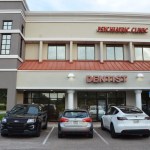 Two-story commercial building with dentistry signage, beige and gray exterior, parking lot with vehicles